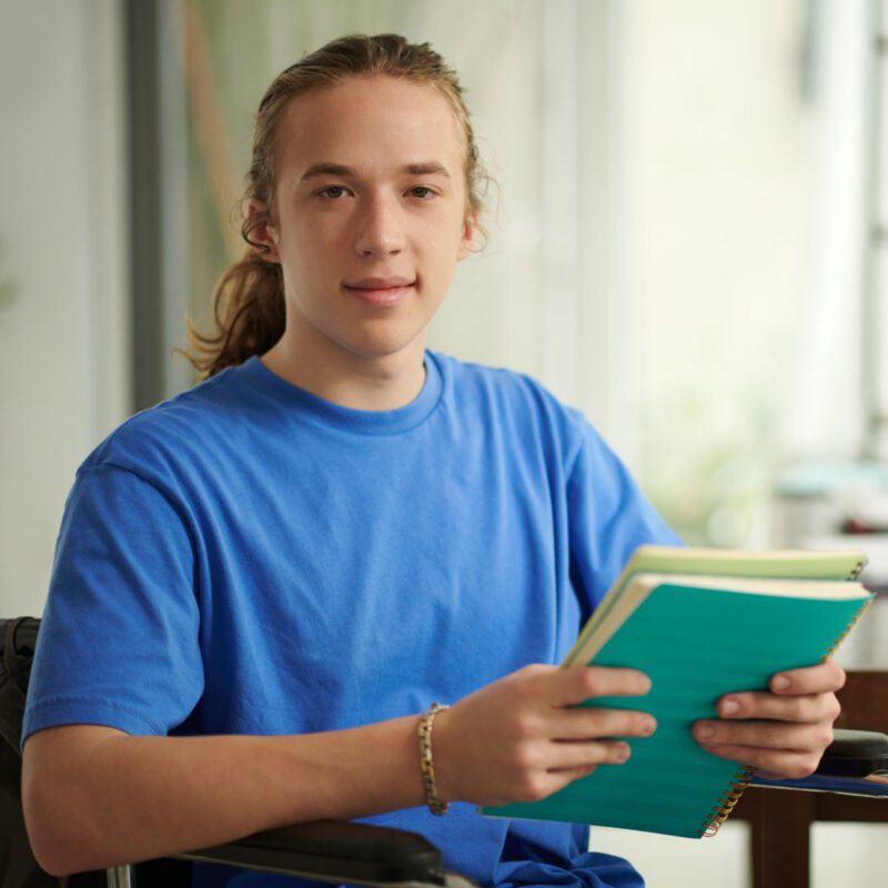 Young adult sitting in a chair holding up a notepad and book.