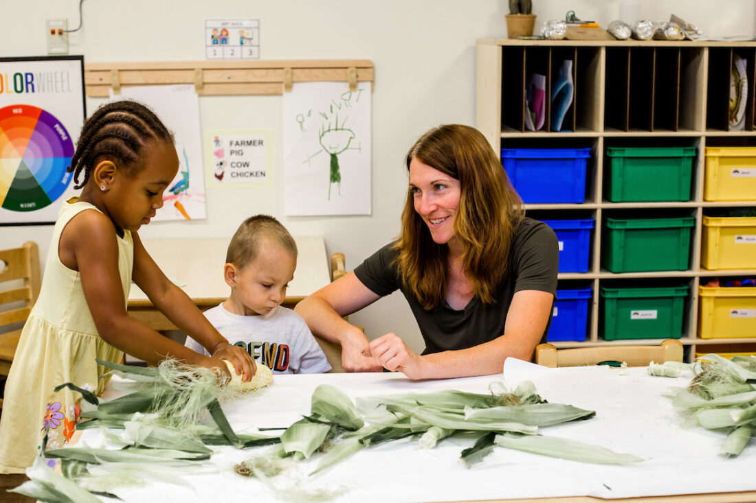 A woman smiling as two children play with corns in a classroom.