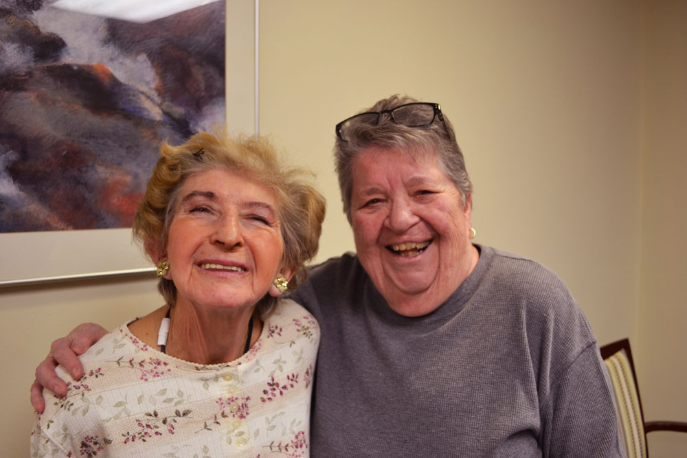 Two old woman standing next to one another smiling brightly at the camera.