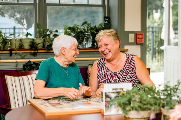 Two old women playing a board game together while smiling brightly at one another.