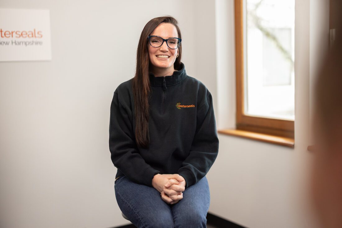 Girl sitting on a wooden stool wearing a black sweatshirt and jeans smiling. 
