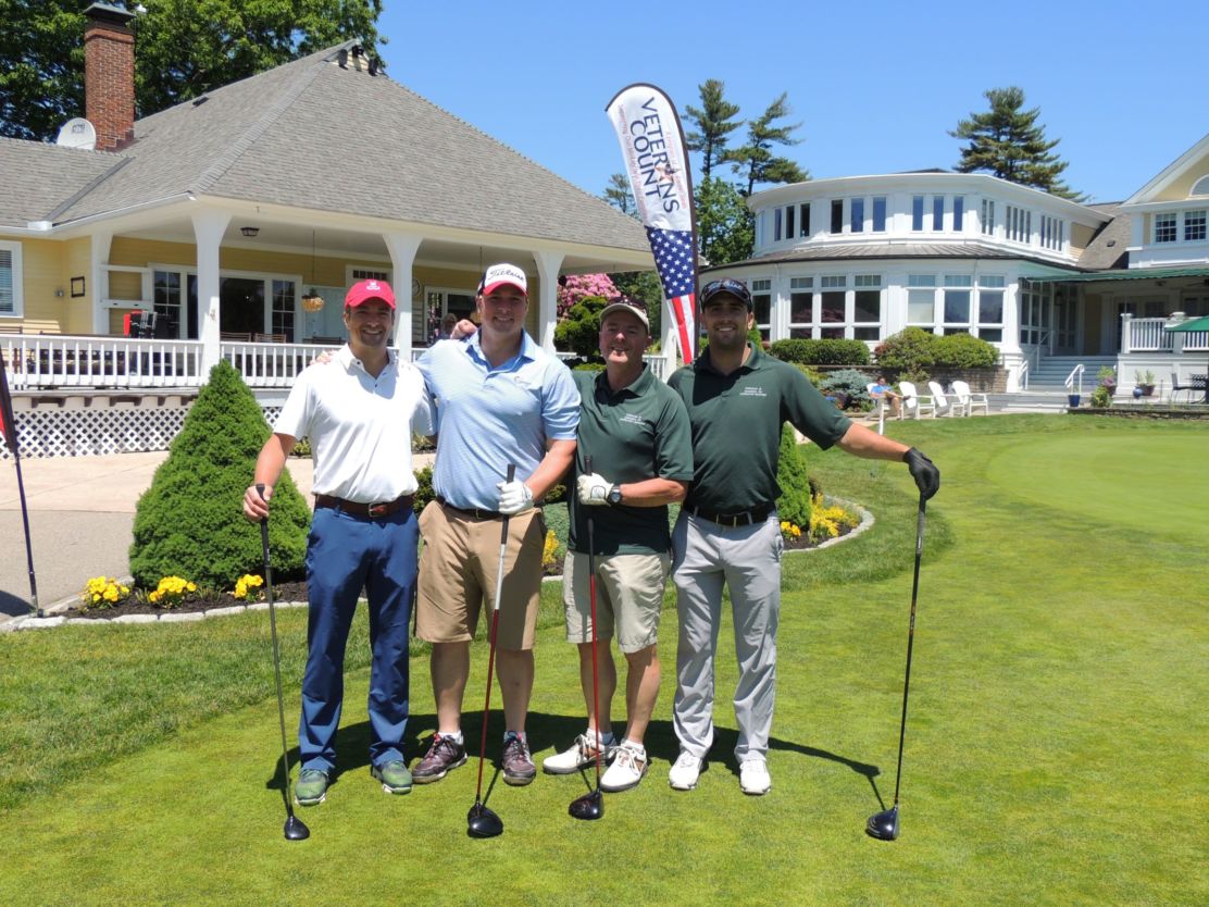 a group of four men with golf clubs stand on a golf green outside a club house