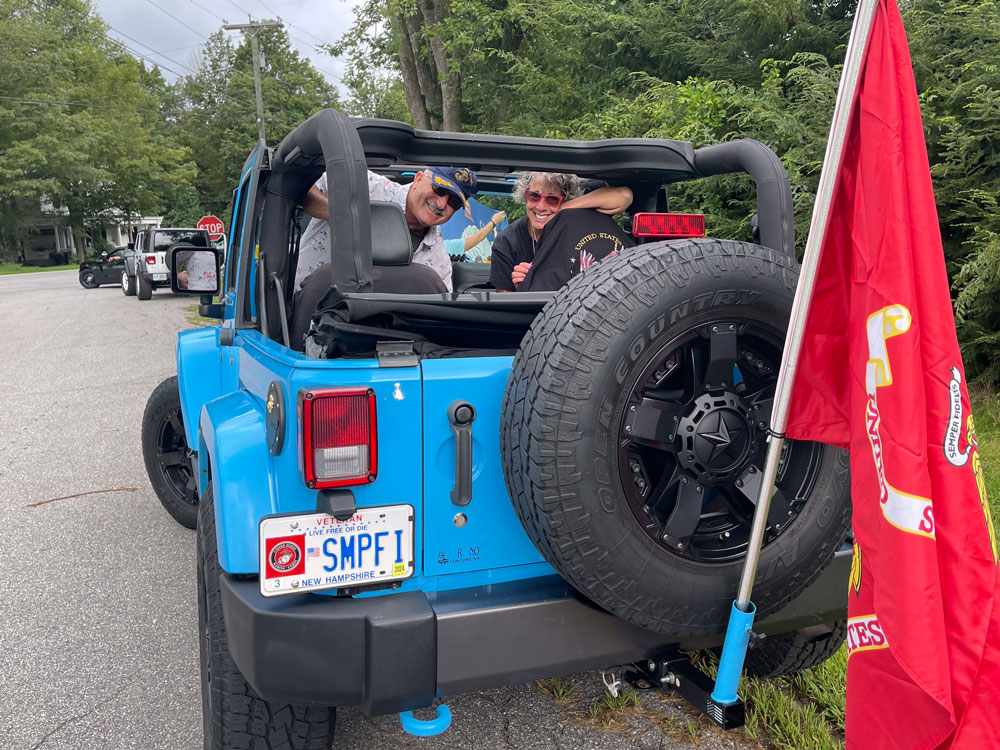 Man and woman inside of a blue jeep with a Marine Corp license plate and Marine Corp flag hanging out the back.